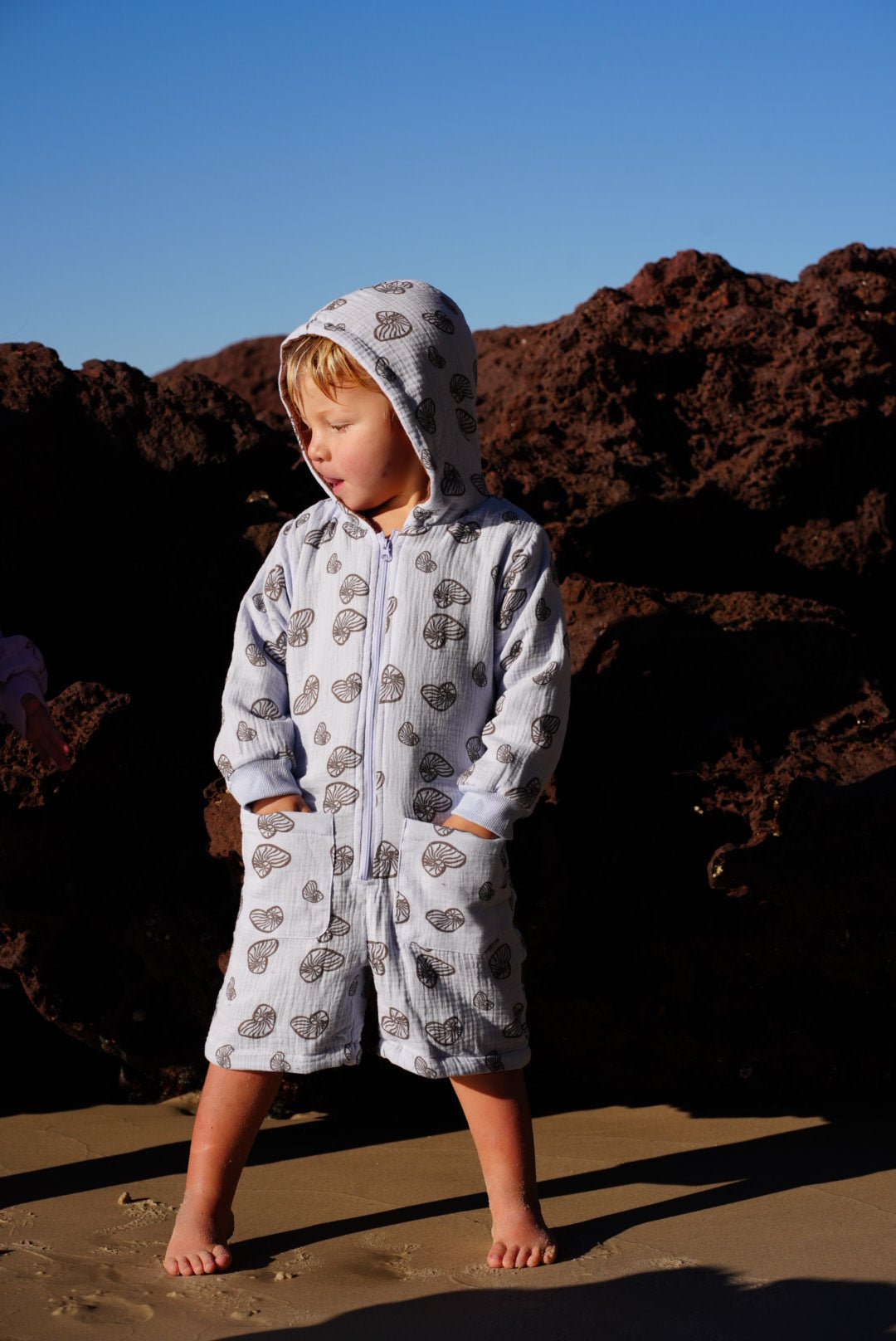 A child wearing a hooded one-piece romper with a light gray and white pattern, standing on a sandy beach next to a dark rock formation under a clear blue sky.