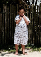 A young child wearing a white short-sleeve romper with a shell print, standing barefoot in front of a rustic wooden fence.