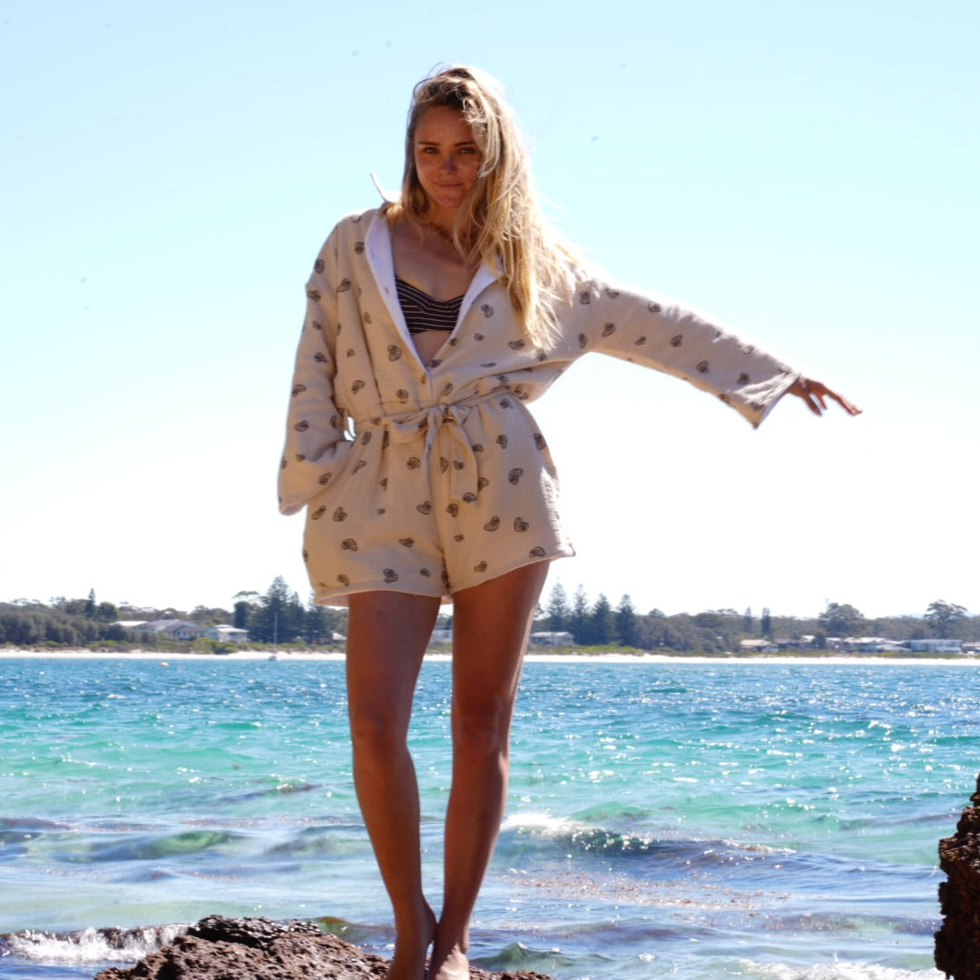 A woman stands on rocks by the ocean wearing a cream-colored, short-sleeved robe with a small brown pattern, over a striped bikini top. The background shows a sunny blue sky and turquoise water.
